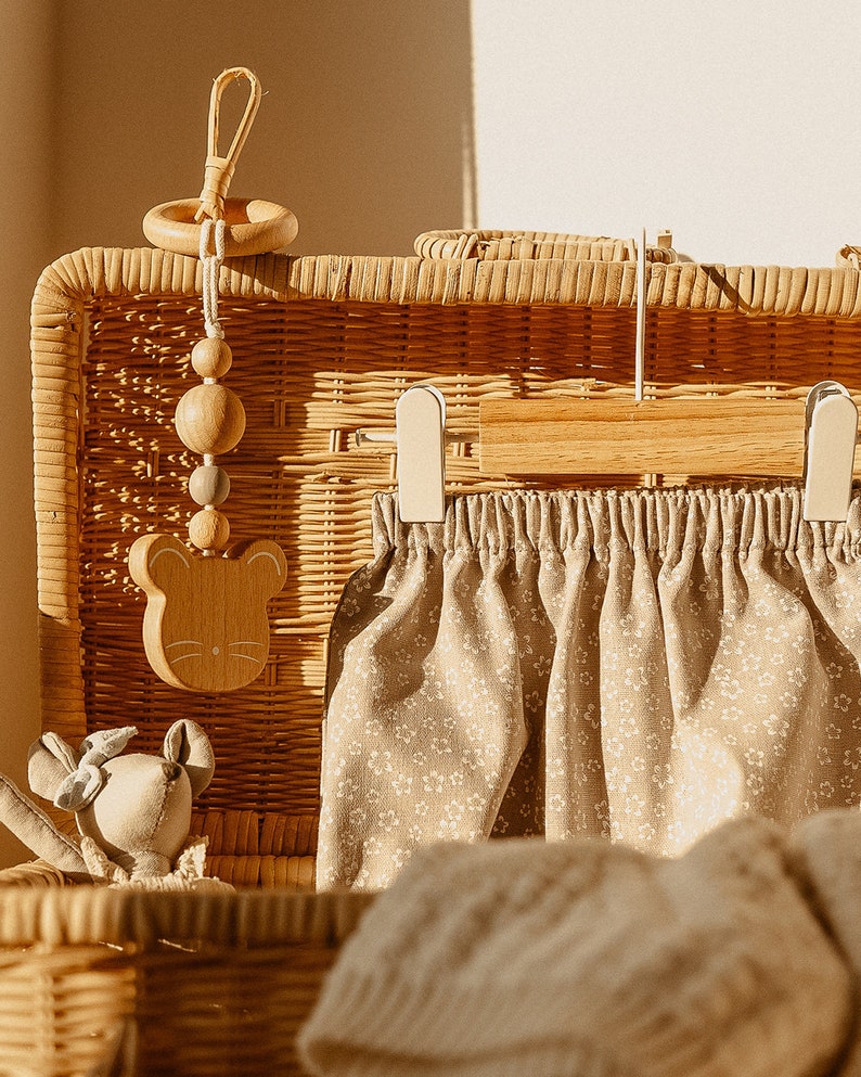 May include: A wicker basket with a wooden toy hanging from the top. The toy is a mouse with a ring and beads. Inside the basket is a pair of beige pants with a floral pattern and a beige knitted blanket.