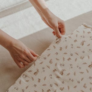 May include: A white muslin swaddle blanket with a delicate pattern of brown bunnies and leaves. The blanket is being folded by a hand.