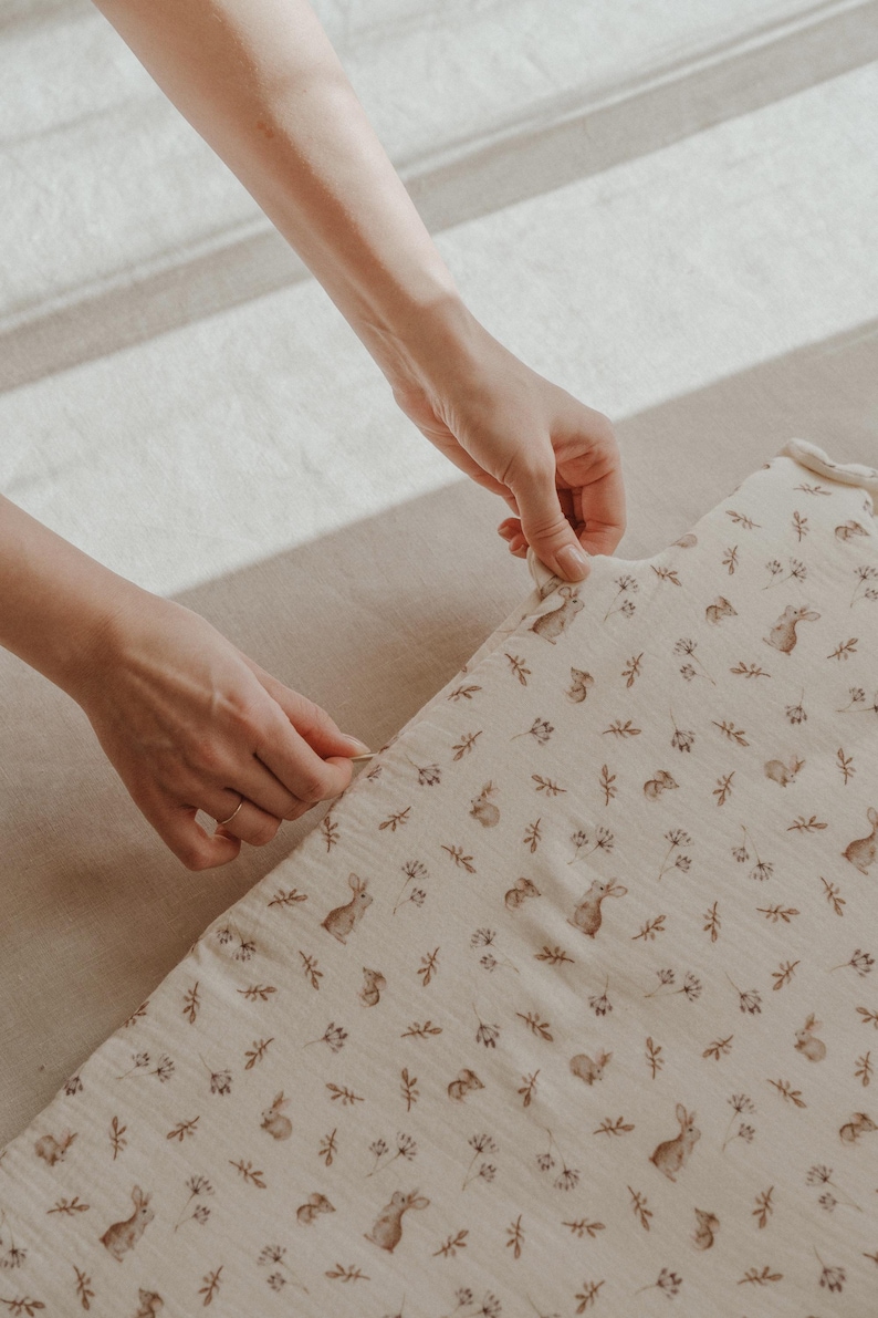 May include: A white muslin swaddle blanket with a delicate pattern of brown bunnies and leaves. The blanket is being folded by a hand.