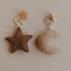 May include: Two plush baby toys, a brown star and a beige crescent moon, each attached to a pacifier clip.