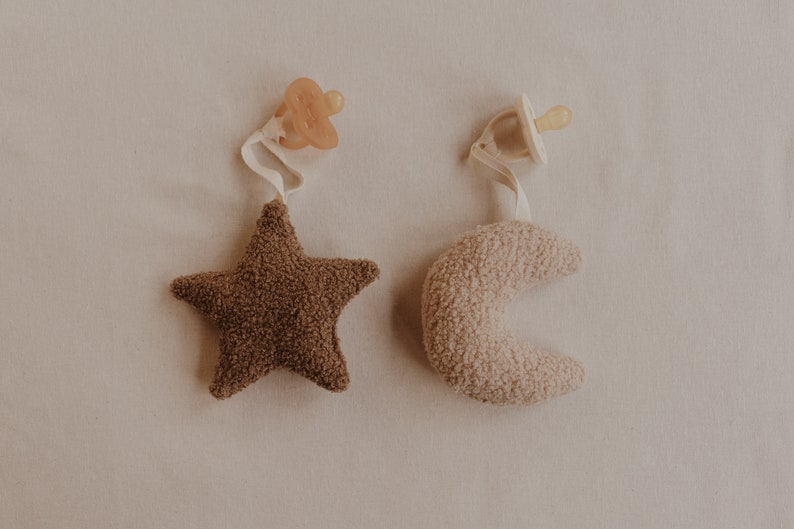 May include: Two plush baby toys, a brown star and a beige crescent moon, each attached to a pacifier clip.