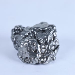 May include: A metallic silver-coloured nugget with an irregular, textured surface featuring various rounded protrusions and deep crevices. The object has a reflective, polished finish that catches the light, creating highlights and shadows across its form.