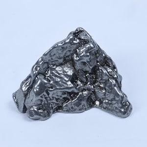 May include: A metallic, dark gray iron meteorite specimen with a highly irregular, pitted, and textured surface. The object features sharp, angular ridges and deep, crater-like indentations across its entire form, showcasing a natural, raw appearance.