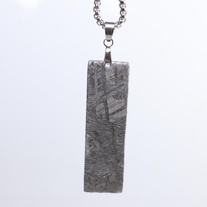 Puede incluir: Un colgante rectangular gris oscuro con un patrón texturizado único, suspendido de una cadena de color plateado. El colgante tiene una apariencia metálica y probablemente está hecho de piedra o metal. El collar se muestra sobre un fondo blanco.