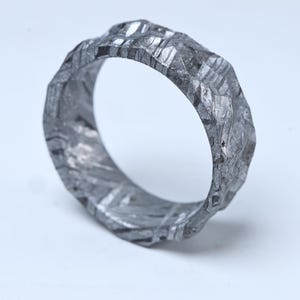 May include: A textured, silver-colored ring with a unique, irregular surface. The ring's design features a pattern of intersecting lines and facets, creating a rugged, yet refined aesthetic. This piece of jewelry is a statement ring.