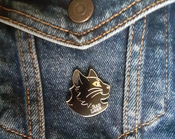 May include: A black enamel pin with a black cat silhouette and the text "Bad Luck" in gold lettering. The pin is attached to a blue denim jacket.