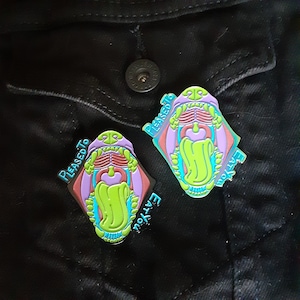 May include: Two enamel pins with a graphic of an open mouth with teeth and a tongue. The pins are blue, green, pink, and purple. The text on the pins reads "Pleased to Eat You".