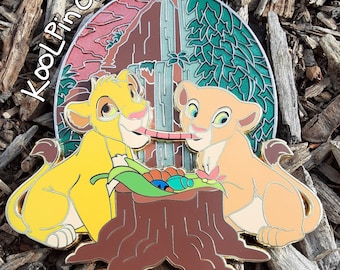 May include: A gold-tone pin featuring Simba and Nala from Disney's The Lion King. The pin depicts the two lion cubs sharing a worm while sitting on a tree stump. The background features a colorful scene of a waterfall and a jungle.