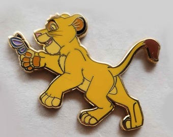 May include: A gold-colored pin featuring a cartoon lion cub with a brown tail, holding a purple and blue butterfly in its paw. The lion cub is walking with its head turned to the side.