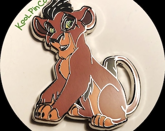 May include: A metal enamel pin featuring a cartoon lion cub with brown fur, black hair, and green eyes. The design includes a light tan muzzle and underbelly, with silver metal outlines. The text KoolPinCo is printed in green in the upper left corner.
