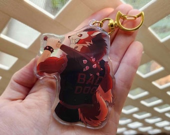 May include: A red and black anime-style wolf character with a baseball bat, wearing a black jacket with the text "BAD DOG" on the back. The character is holding the bat in its right paw and has a fierce expression. The character is a keychain made of clear acrylic.