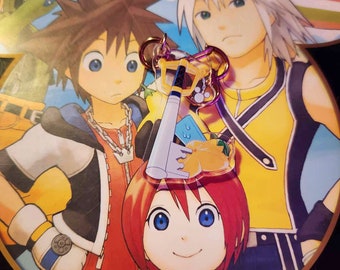 May include: A colorful keyblade charm with a yellow flower, blue and white details, and a gold chain. The charm is held by a person with red hair and blue eyes. The background features cartoon characters.