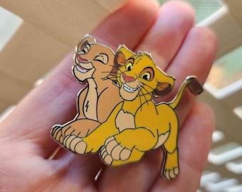 May include: A metal enamel pin featuring two cartoon lion cubs. One cub is yellow with a dark tail tip, and the other is light tan. Both cubs have detailed facial features, including black whiskers, pink noses, and expressive eyes, rendered in a classic animation style with a silver-toned metal outline.
