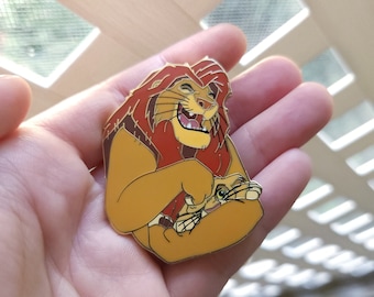 May include: A gold-colored enamel pin featuring Simba, the lion from Disney's The Lion King, with a playful expression. Simba is holding a smaller lion cub, with a brown and tan color scheme.