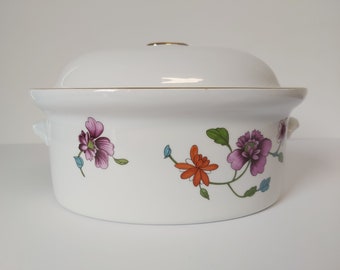 Op de afbeelding: Witte ovale keramische ovenschaal met deksel. De schaal heeft een gouden rand en een bloemenmotief met paarse, oranje en groene bloemen.
