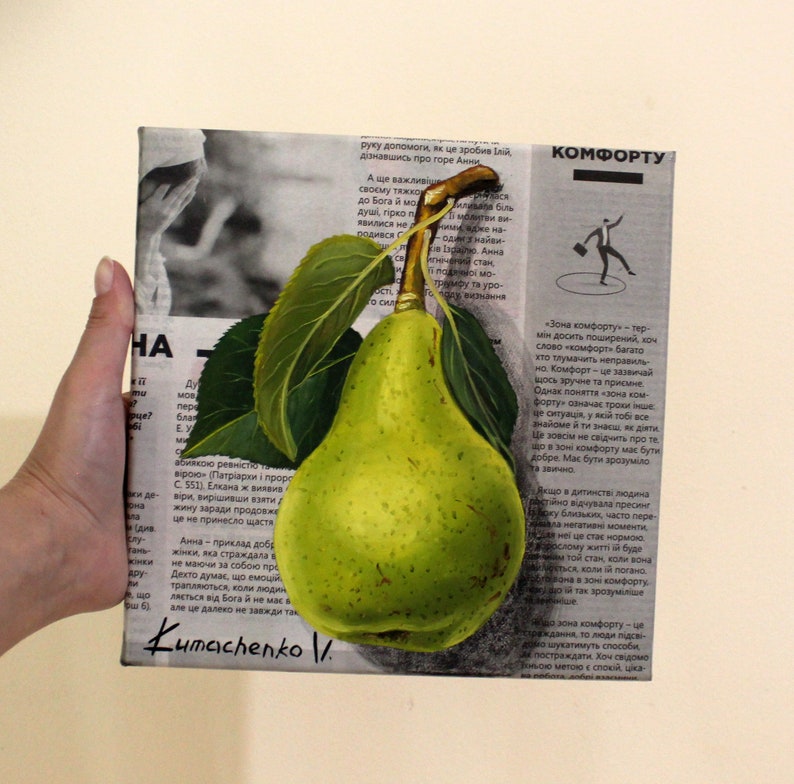 May include: A square canvas painting featuring a realistic, bright green pear with two dark green leaves and a brown stem. The pear is centered over a background of black and white printed newspaper text in Ukrainian, with the signature Kumachenko V. at the bottom.