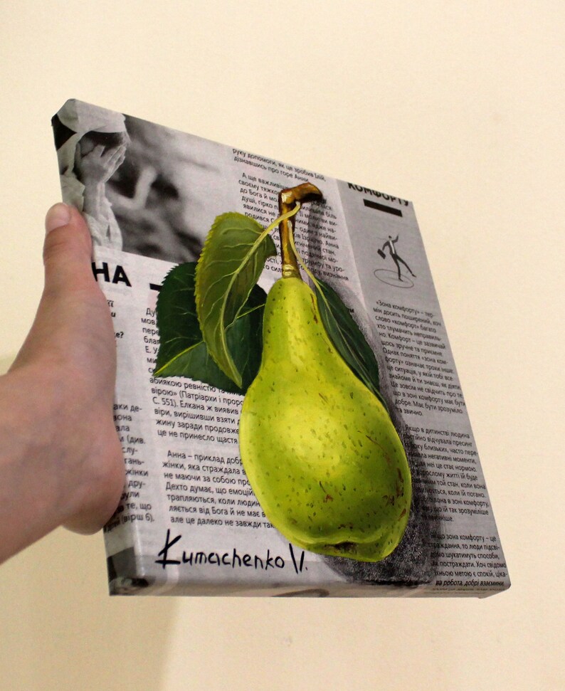 May include: A square canvas print featuring a realistic painting of a bright green pear with two dark green leaves attached to its stem. The pear is superimposed over a black and white background of printed newspaper text and a small photograph.