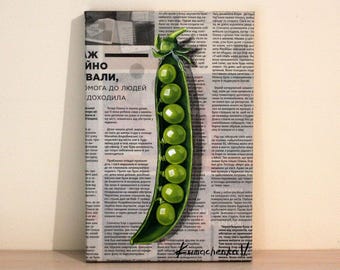 May include: A painting of a green pea pod with a stem and leaves. The pod is filled with green peas. The painting is on a canvas with a newspaper background. The text on the newspaper is in Russian.