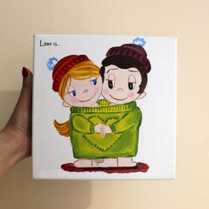 Könnte beinhalten: Leinwandgemälde mit zwei Cartoonfiguren, die sich umarmen und einen grünen Pullover tragen. Eine Figur hat blondes Haar und eine rote Mütze, die andere hat dunkles Haar und eine kastanienbraune Mütze. Der Text "Love is..." steht oben links.