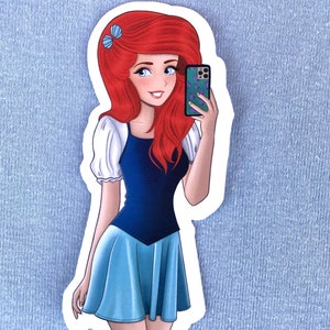 May include: A sticker featuring a cartoon illustration of a character with long red hair, wearing a blue dress and holding a phone. The phone has a green case with a purple design. The character is taking a selfie.