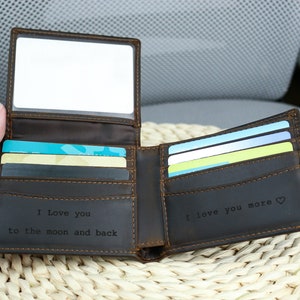 May include: A brown leather wallet with a clear plastic window on the front. The wallet is open and has several cards inside. The inside of the wallet is engraved with the text 'I love you to the moon and back' and 'I love you more'. The wallet is sitting on a woven wicker surface.