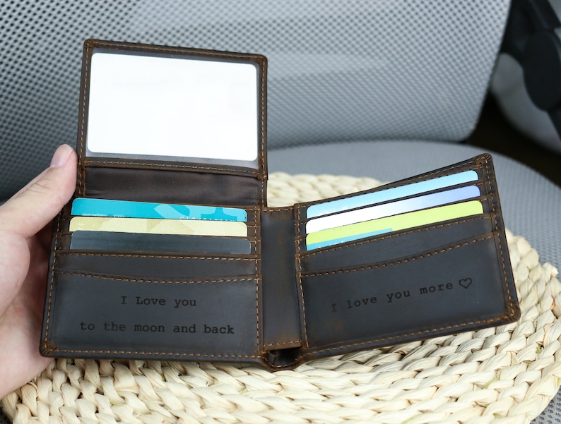 May include: A brown leather wallet with a clear plastic window on the front. The wallet is open and has several cards inside. The inside of the wallet is engraved with the text 'I love you to the moon and back' and 'I love you more'. The wallet is sitting on a woven wicker surface.