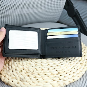 May include: A black leather wallet with a clear plastic window for a photo ID. The wallet is open and has several card slots and a section for cash. The wallet is engraved with the text 'Happy Birthday Dad 12.11.2021'.