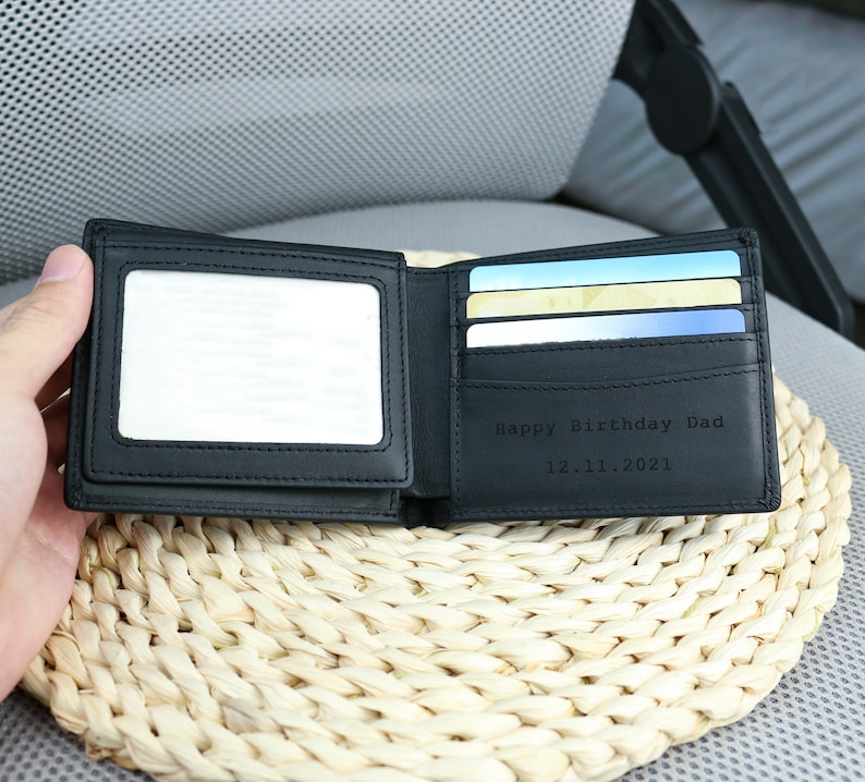 May include: A black leather wallet with a clear plastic window for a photo ID. The wallet is open and has several card slots and a section for cash. The wallet is engraved with the text 'Happy Birthday Dad 12.11.2021'.