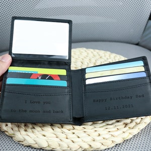 May include: A black leather wallet with a mirror on the inside. The wallet has a card slot and is engraved with the text 'I love you to the moon and back' on one side and 'Happy Birthday Dad 12.11.2021' on the other side.