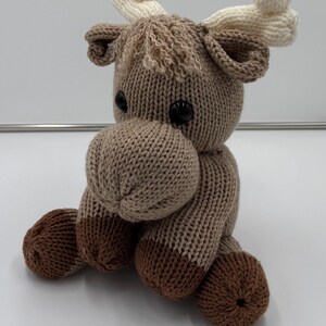 May include: A knitted moose toy in brown and beige. The moose has black button eyes, a rounded snout, and cream-coloured antlers. The toy is sitting with its legs bent, showing its brown hooves. A charming handmade item.