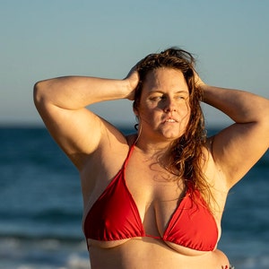 May include: A woman with long, wet hair is wearing a red bikini top while standing on a beach. The ocean is in the background.