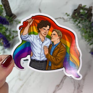 May include: A die-cut sticker featuring an illustration of two men standing together under a large rainbow flag. One man has dark curly hair and wears a white button-down shirt, while the other has light hair and wears a brown jacket over a blue shirt.