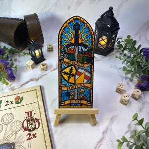 Può includere: Un'opera d'arte in stile vetrata raffigurante una spada, fiori e scudi, esposta su un piccolo cavalletto di legno. L'opera d'arte include le parole "AUDENTES" e "FORTUNA IUVAT". Lo sfondo è blu e giallo.