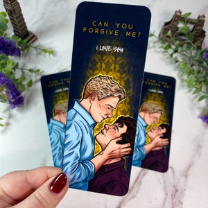 May include: Bookmarks with a romantic illustration of two men, one in a blue shirt and the other in a purple shirt, with the text "CAN YOU FORGIVE ME? I LOVE YOU" on a navy blue background with a gold pattern.