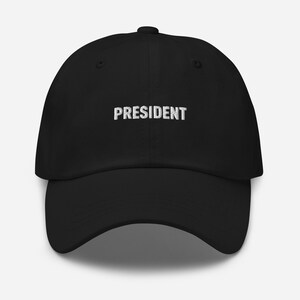 May include: Black baseball cap with the word "PRESIDENT" embroidered in white across the front. The cap has a curved brim and a button on top. A classic, casual accessory.