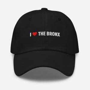 Puede incluir: Gorra de béisbol negra con el texto bordado "I ❤️ THE BRONX". El corazón es rojo y el texto es blanco. La gorra tiene una visera curva y una correa ajustable.