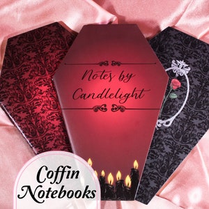 May include: Three black and red coffin-shaped notebooks with a damask pattern. The centre notebook has the text "Notes by Candlelight" in black script. The notebooks are on a pink satin background.