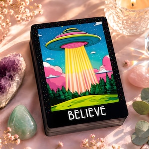May include: A rectangular deck of cards featuring a colorful illustration of a flying saucer hovering over a forest and emitting a bright yellow beam of light. The card has a black border and the word BELIEVE printed in white at the bottom.