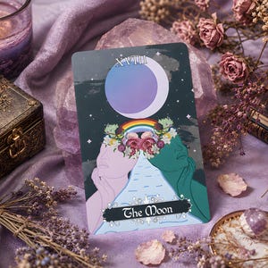 Puede incluir: Una carta de tarot rectangular titulada The Moon con el número romano XVIII, una gran luna creciente morada, un arcoíris y dos rostros de perfil en rosa claro y verde azulado. La carta muestra un arreglo floral central y un diseño de agua que fluye.