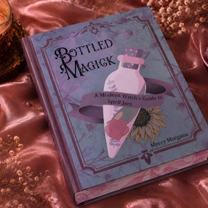May include: A hardback book titled Bottled Magick: A Modern Witch's Guide to Spell Jars by Mercy Morgana. The cover features a light purple background with a central illustration of a glass bottle containing a pink rose, a sunflower, and lavender.