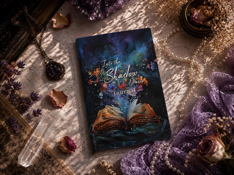 Puede incluir: Un diario rectangular con una cubierta azul oscuro que presenta una ilustraci&oacute;n gr&aacute;fica de un libro abierto que emite flores coloridas y luz. El texto en la cubierta dice Into the Shadow Oracle Journal en fuentes blancas y azul claro.