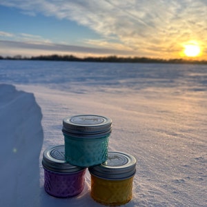 May include: Three small, colorful jars with silver lids are stacked on a snowy surface. The jars are teal, purple, and yellow. The background features a winter landscape with a sunset and a blue sky.