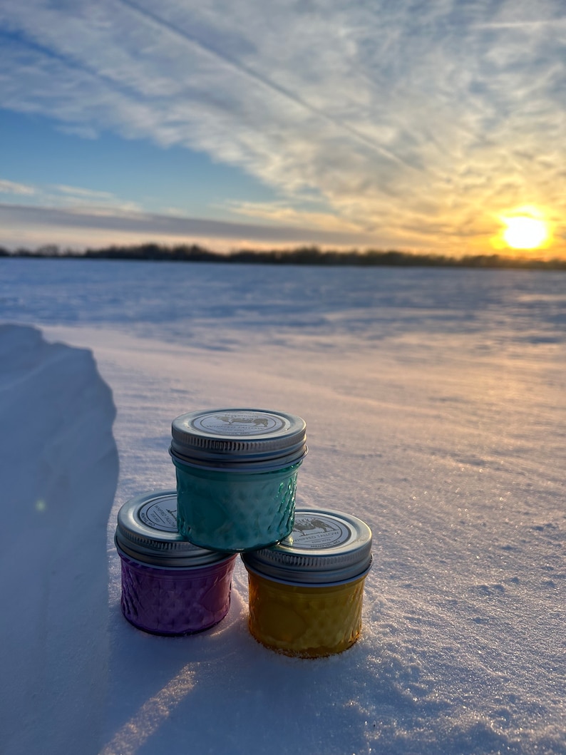 May include: Three small, colorful jars with silver lids are stacked on a snowy surface. The jars are teal, purple, and yellow. The background features a winter landscape with a sunset and a blue sky.