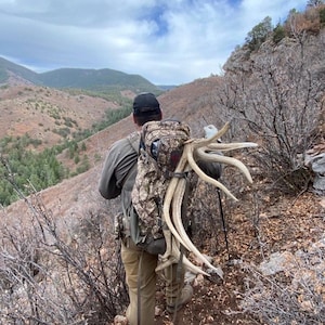 May include: A person hiking in a mountainous area, carrying a camouflage backpack and large antlers. The landscape features brown and green vegetation under a cloudy sky. The person is wearing a black cap and khaki pants.