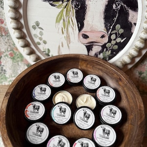 May include: A wooden bowl filled with small, round tins of whipped tallow. Each tin has a black lid with a white label featuring a cow illustration and the text "WHIPPED TALLOW". Some tins are open, revealing the creamy product.