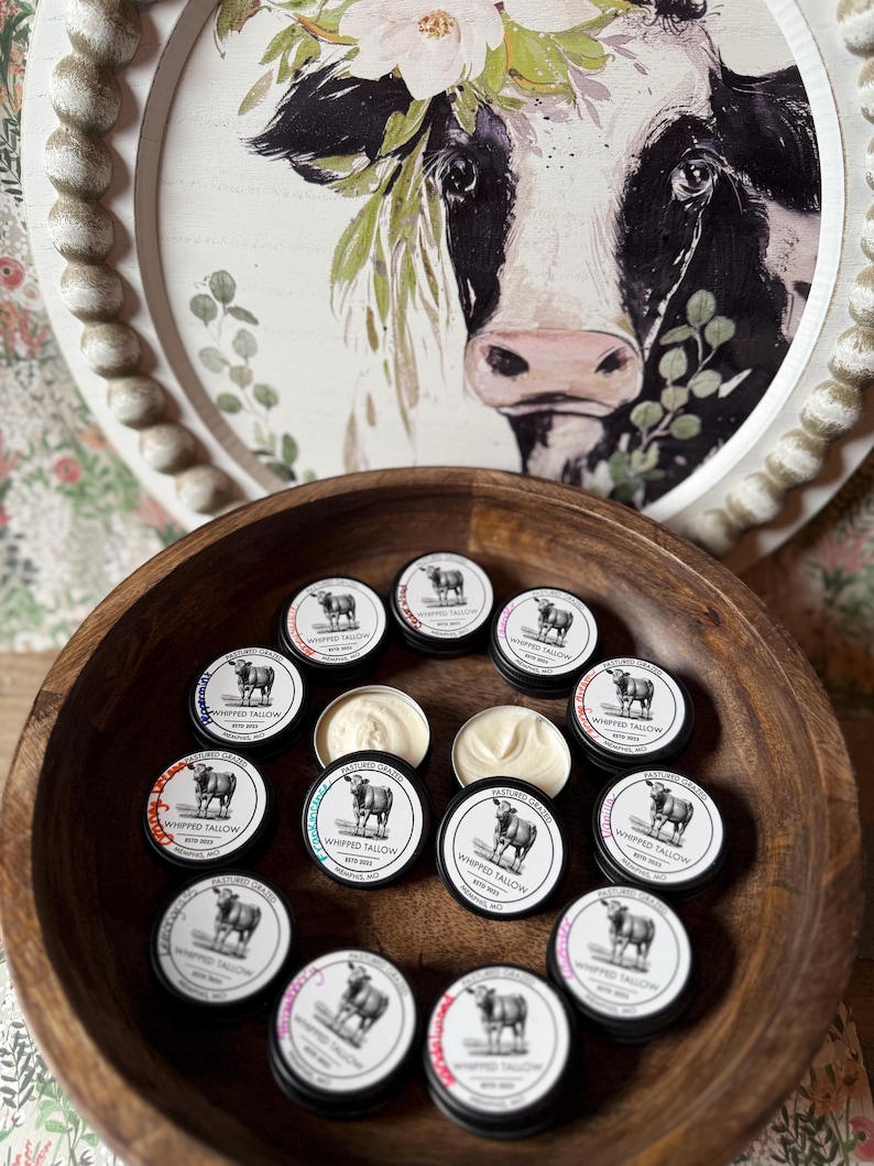 May include: A wooden bowl filled with small, round tins of whipped tallow. Each tin has a black lid with a white label featuring a cow illustration and the text "WHIPPED TALLOW". Some tins are open, revealing the creamy product.