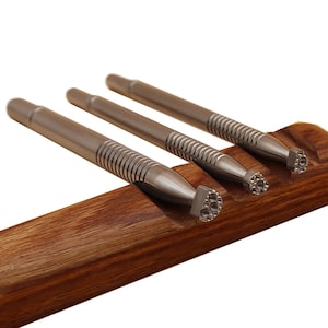 Op de afbeelding: Drie zilveren metalen stempelgereedschappen met verschillende gepatroneerde uiteinden worden getoond in een houten houder. De gereedschappen hebben een getextureerde grip en worden gebruikt voor het verwerken van leer.