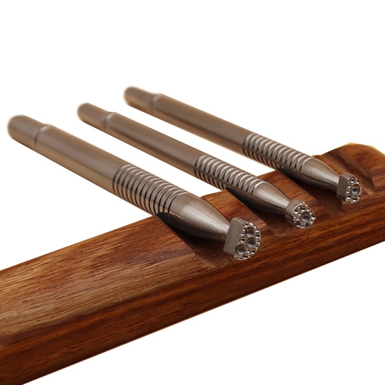Op de afbeelding: Drie zilveren metalen stempelgereedschappen met verschillende gepatroneerde uiteinden worden getoond in een houten houder. De gereedschappen hebben een getextureerde grip en worden gebruikt voor het verwerken van leer.
