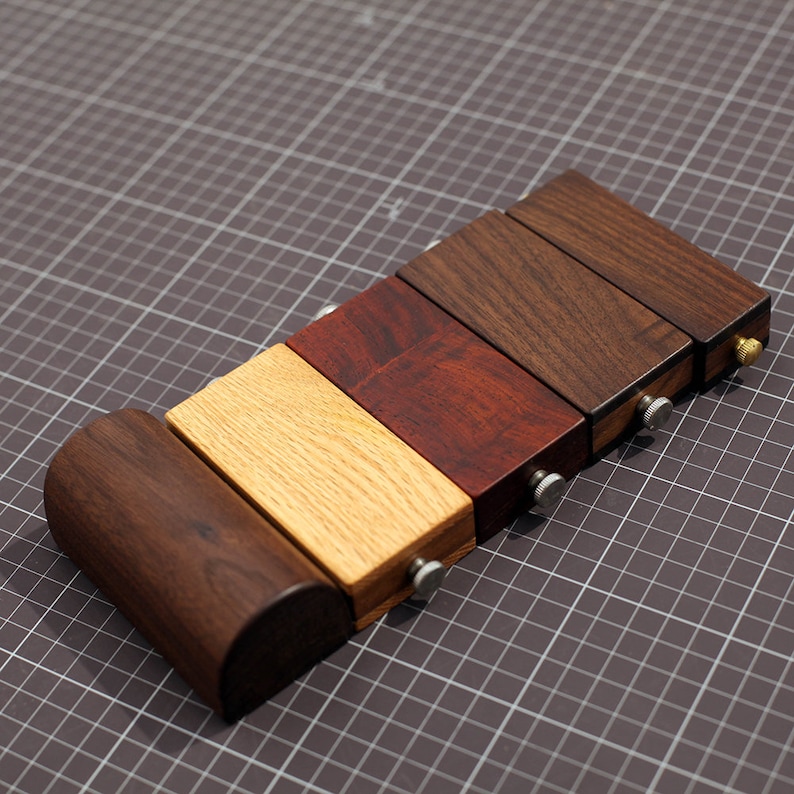 May include: Five wooden blocks of different colors and wood grains, stacked on top of each other. The blocks are connected with metal screws. The blocks are on a gray grid background.