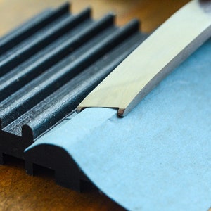 May include: A close-up of a metal tool being used to cut a piece of blue paper. The tool has a curved edge and is being held at an angle. The paper is lying on a black and gray textured surface.